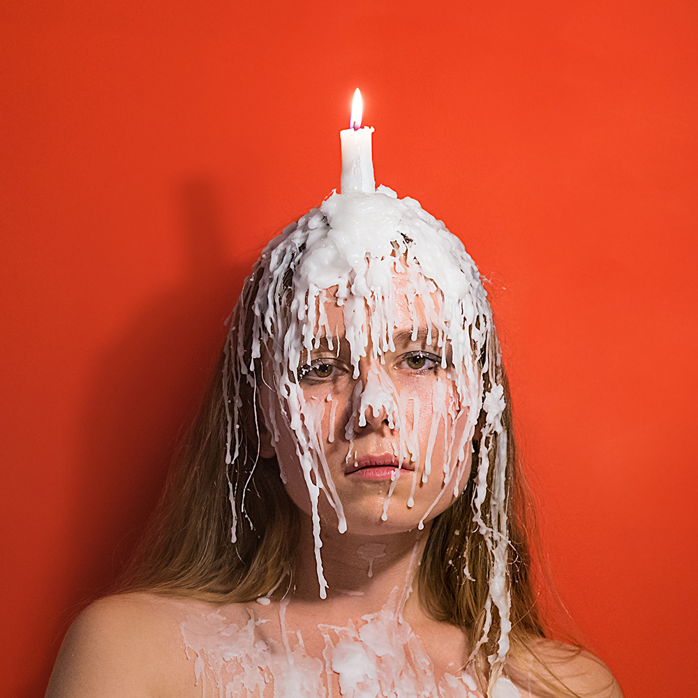 Headshot of Sage Szkabarnicki-Stuart with a candle on her head, dripping wax on her face and chest, in front of an orange background.